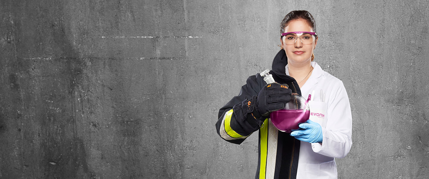 Fotomontage einer Frau: Zur einen Hälfte in weißem Laborkittel, zur anderen in Feuerwehruniform. Sie hält einen Rundkolben mit einer farbigen Flüssigkeit in den Händen.