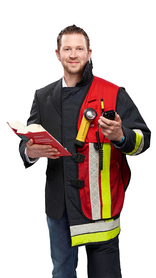 Fotomontage eines Mannes: Halb Verwaltungswirt in dunklem Sakko und Jeans, halb Feuerwehrmann in Uniform. Er trägt ein Funkgerät, eine rote Gruppenführerweste und ein Buch mit Gesetzestexten.