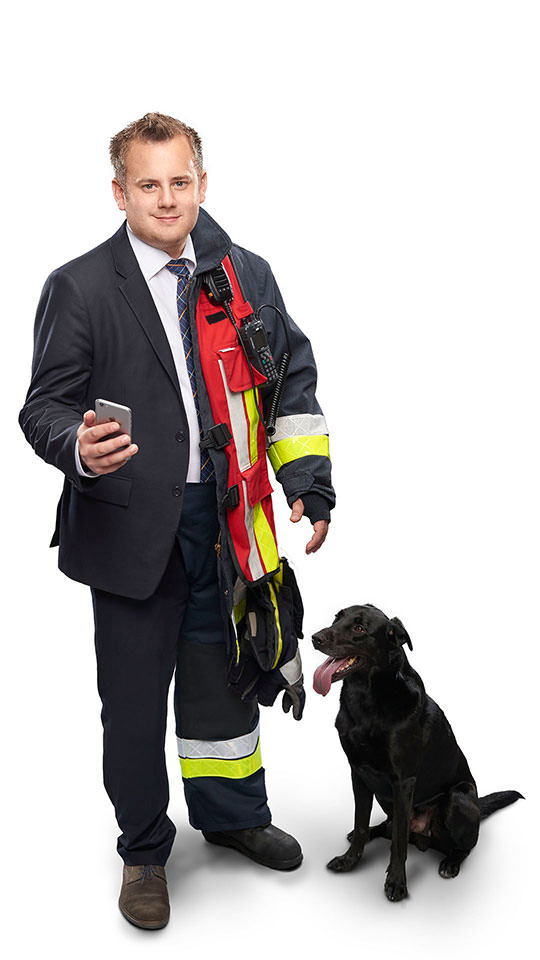 Fotomontage eines Mannes: Zur einen Hälfte im Businessanzug mit Handy, halb Feuerwehrmann in Uniform. Neben ihm sitzt ein schwarzer Hund.