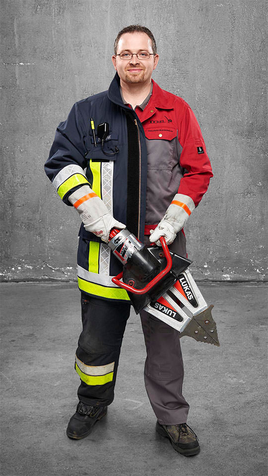 Eine Fotomontage eines Mannes: Halb in roter Arbeitskleidung, halb Feuerwehrmann in Uniform. Er hält ein hydraulisches Arbeitsgerät vor sich.