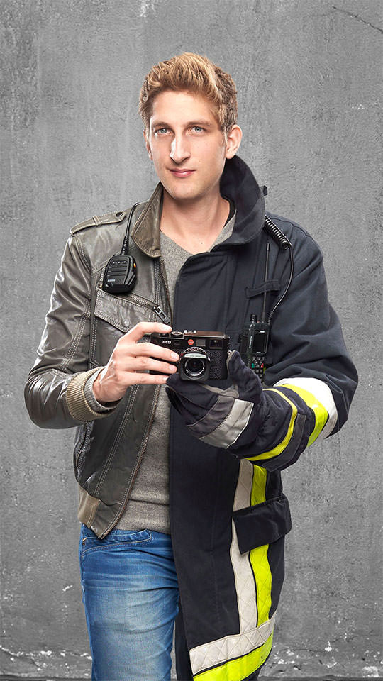 Fotomontage eines Mannes: Zur einen Hälfte mit grüner Lederjacke und grauem Pullover, zur anderen in Feuerwehruniform. Er hält eine Kamera in den Händen.