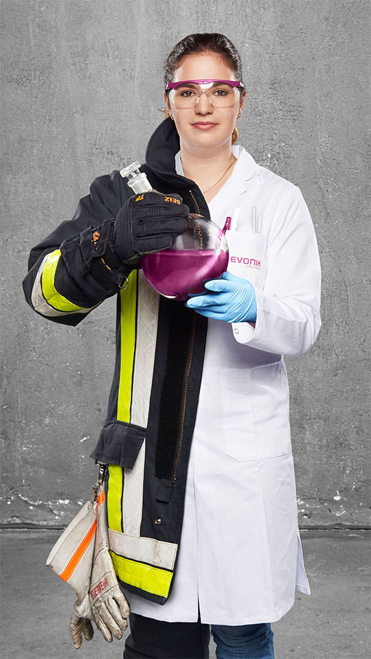 Fotomontage einer Frau: Zur einen Hälfte in weißem Laborkittel, zur anderen in Feuerwehruniform. Sie hält einen Rundkolben mit einer farbigen Flüssigkeit in den Händen.